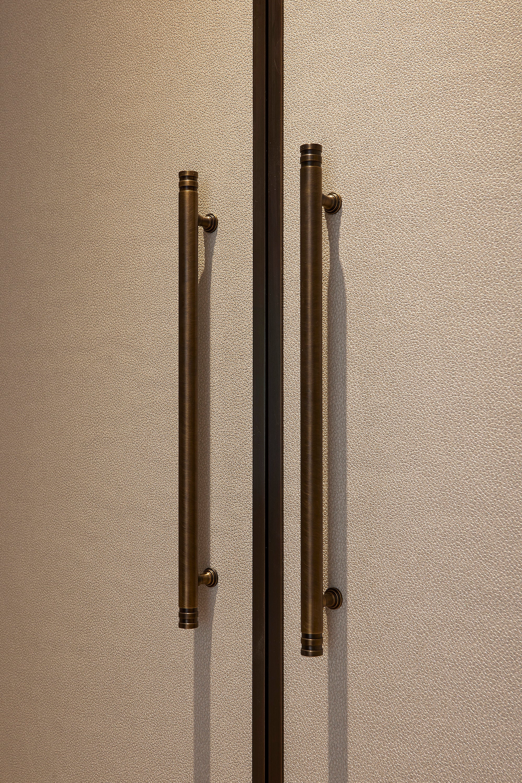 Close-up of a door with a pair of antique brass 'henley' cabinet pull handles on a beige leather cladded door.